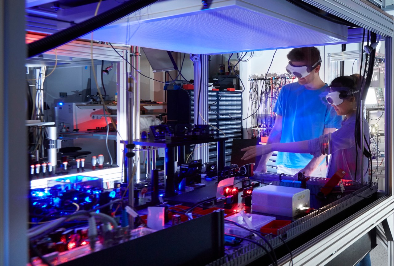 Scientists standing next to a laser table-top radiating purple light in an quantum simulation lab. 