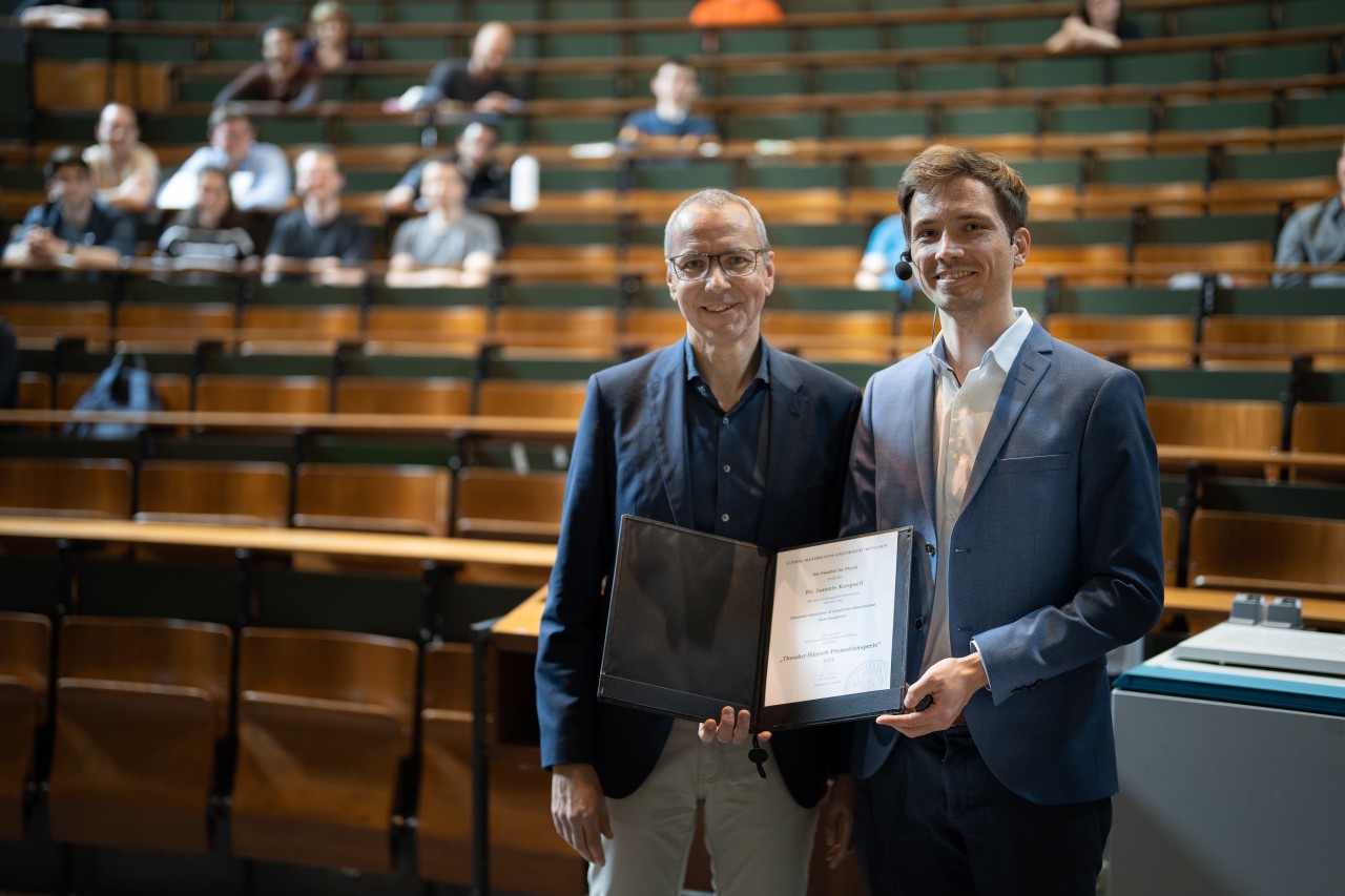 Two men standing next to eachother holding a diploma in a lecture hall.